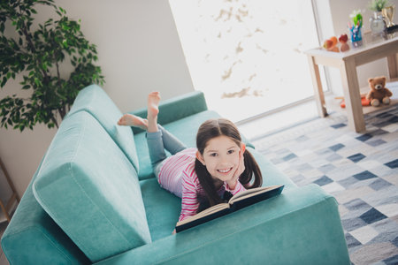 Photo portrait of young funny kid little brunette girl staying at home spending hobby time lying couch enjoying her book isolated in her roomの写真素材