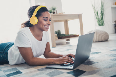 Portrait of nice young woman lay floor use laptop listen music wear t-shirt modern interior flat indoorsの写真素材