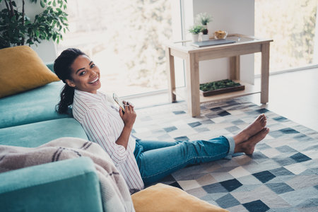 Full body portrait of nice young woman sit floor hold smart phone wear shirt modern interior flat indoorsの写真素材