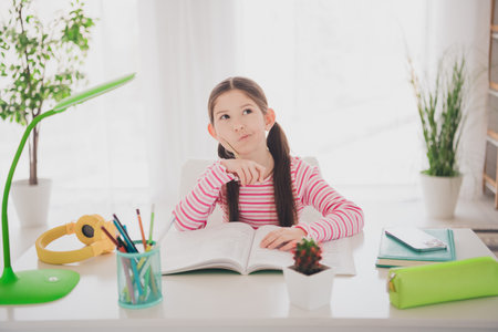 Photo of young brunette schoogirl staying at home deep thinking preparing for exam reading book sitting desk holidays isolated in her roomの写真素材