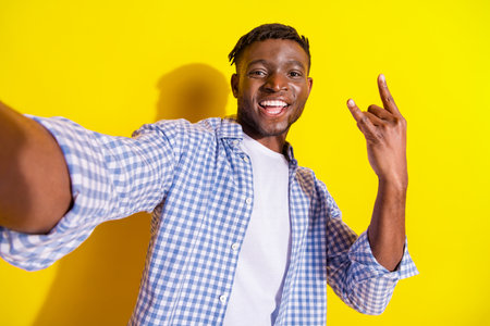 Photo of cheerful positive guy dressed checkered shirt recording video showing hard rock sing emtpy space isolated yellow color backgroundの写真素材
