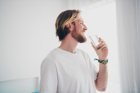 Photo of attractive young male drink water glass morning dressed sleepwear cozy day light home interior living roomの写真素材