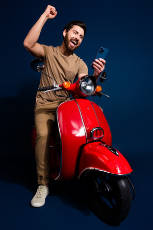 Vertical photo of overjoyed man in beige t shirt riding red vintage motorbike raised fist up using phone isolated on dark blue color backgroundの写真素材