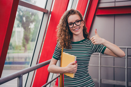 Portrait of nice young girl striped dress hold book show thumb up work study office business centerの写真素材