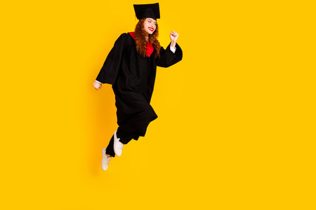 Photo of cute positive girl student wear black mantle jump up run fast isolated on yellow color backgroundの写真素材