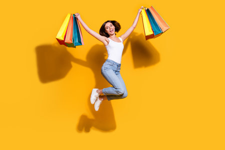Full body photo of attractive young girl hold shopping bags jumping excited wear trendy white outfit isolated on yellow color backgroundの写真素材