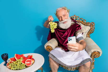 Portrait of ancient myth god smart phone make selfie hold grape sit throne isolated on blue color backgroundの写真素材