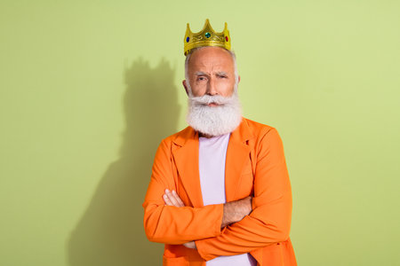 Portrait of aged man performer golden crown crossed arms wear orange suit isolated on green color backgroundの写真素材