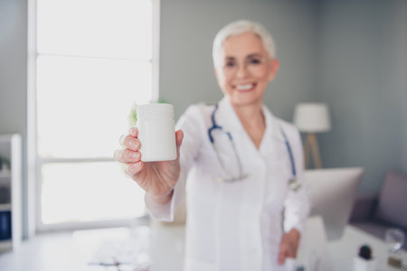 Portrait of aged woman professional doctor hold pills jar wear white coat workplace office indoorsの写真素材