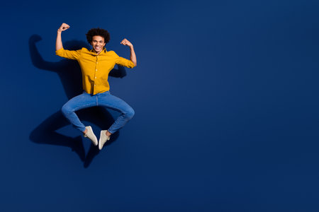 Full body portrait of nice young man jump raise fists flex biceps wear yellow shirt isolated on dark...