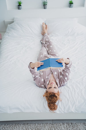 Full body vertical portrait of pretty young girl read book comfy bed wear pajama bedroom apartment indoorsの写真素材