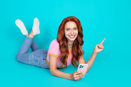 Full body photo of attractive young woman lying floor hold device dressed stylish pink clothes isolated on cyan color backgroundの写真素材