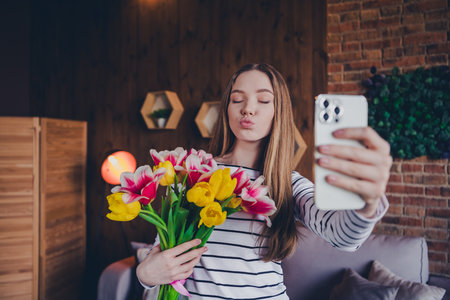 Photo of lovely teen lady hold tulips bouquet selfie photo air kiss dressed striped garment spend pastime spacious house indoors roomの写真素材