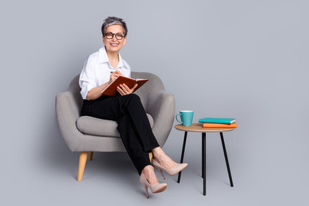 Full body photo of attractive pensioner woman sit armchair write notebook dressed stylish formalwear isolated on gray color backgroundの写真素材