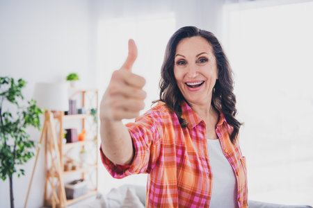 Photo of shiny cute woman dressed checkered shirt showing you thumb up indoors house apartment roomの写真素材