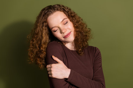 Photo of charming positive girl wear brown shirt cuddling herself empty space isolated green color backgroundの写真素材