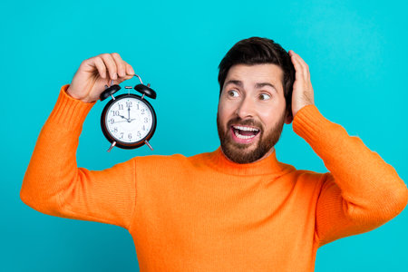 Photo of shocked young man wear trendy clothes hand hold alarm clock isolated on aquamarine color...