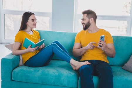 Portrait of two young people hold book show smart phone sofa weekend pastime modern interior flat indoorsの写真素材