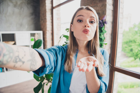 Selfie photo of young homey woman in blue shirt blonde hair realtor smm manager recording video real estate review and sending kisses indoorsの写真素材