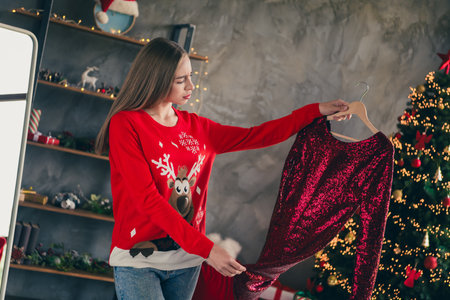 Photo of attractive young woman unsure choose dress wear red christmas outfit cozy day light home party decoration interior living roomの写真素材