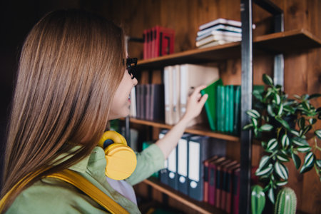 Photo of positive good mood lady student dressed shirt earphones choosing book indoors classroomの写真素材