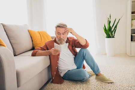 Full length photo of nice pensioner man sit floor confused read instruction dressed brown garment...