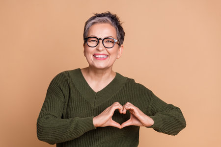 Photo of kind appreciative woman dressed khaki sweater in glasses showing heart symbol on chest isolated on beige color backgroundの写真素材