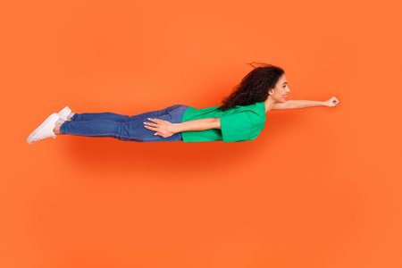 Photo of young attractive american girl wearing green casual t shirt levitating like superhero fist up isolated on orange color backgroundの写真素材