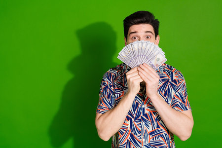 Photo of astonished shy guy with stylish haircut dressed print shirt hiding face behind dollars isolated on green color backgroundの写真素材