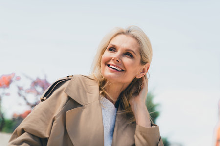 Portrait of attractive senior dreamy female resting strolling enjoy weekend touch hair feminine wear beige coat pastime fresh air outdoorの写真素材