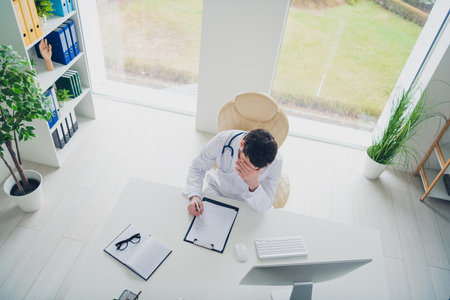 Photo of troubled doctor sit table hide face suffer write clipboard in hopstal clinicの写真素材