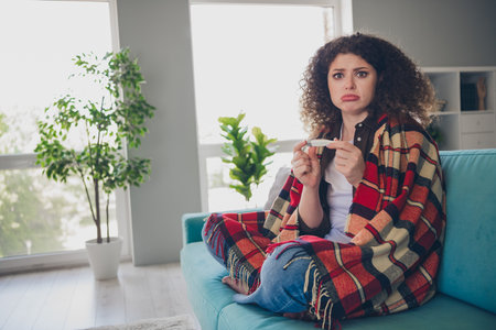 Photo portrait of curly haired young girl sitting comfortable couch wrapped checkered blanket sick holding thermometer crying upsetの写真素材