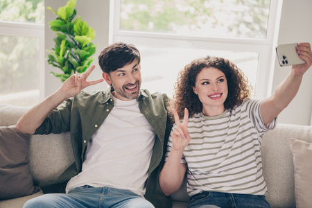 Photo of two young people sit couch smart phone selfie v-sign modern interior apartment indoors