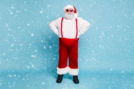 Fulle length photo of funny funky fat overweight grey-haired santa claus with hat modern spectacles enjoy christmas time wear handsome overalls red pants boots isolated over blue color backgroundの写真素材