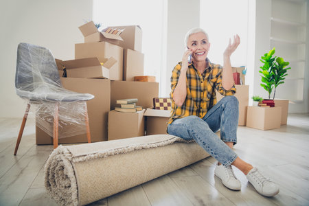 Full size photo of nice aged woman sit rolled carpet communicate phone wear checkered shirt relocate...