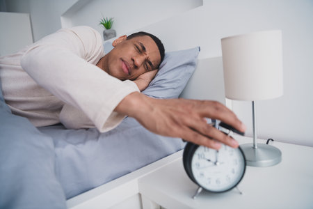 Photo of nice young man lying bed wake up alarm clock wear pajama enjoy wake up modern cozy interior bedroom indoorsの写真素材