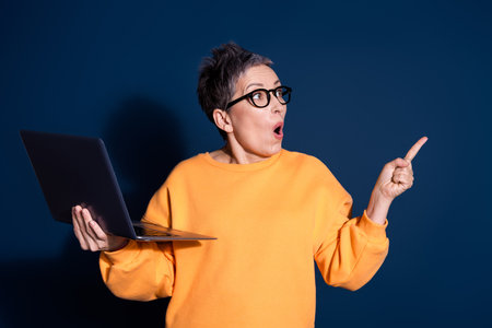 Photo of attractive mature woman hold netbook point look empty space dressed stylish yellow clothes isolated on dark blue color backgroundの写真素材