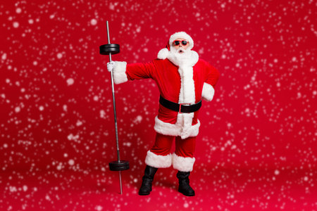 Boss of gym. Full length photo of cool retired old man white beard hold barbell shock light weight wear santa x-mas costume coat sunglass headwear leather boots isolated red color backgroundの写真素材