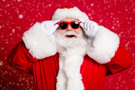 Cant believe my eyes. Closeup portrait photo of retired old man grey beard open mouth smiling hold glasses see north pole wear santa costume sunglass headwear isolated red color backgroundの写真素材