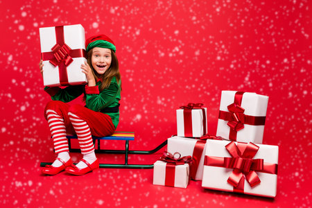 Nice attractive cheerful cheery glad excited funny pre-teen elf sitting on sled holding in hands fairy miracle tradition purchase wish wintertime isolated over bright vivid shine red backgroundの写真素材