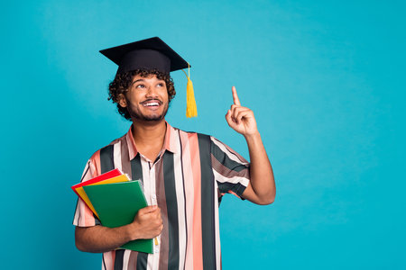 Photo of cheerful celever guy dressed striped shirt mortarboard holding copybooks showing finger up emtpy space isolated blue color backgroundの写真素材