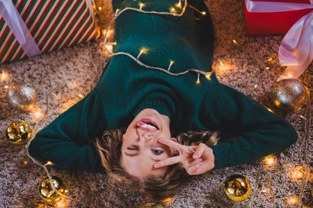 Photo of attractive teen girl lying floor relax v-sign dressed green christmas outfit day light home party decoration interior living roomの写真素材