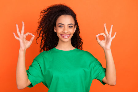 Photo of young funky attractive american girl wearing green casual t shirt and showing two okey sign isolated on orange color backgroundの写真素材