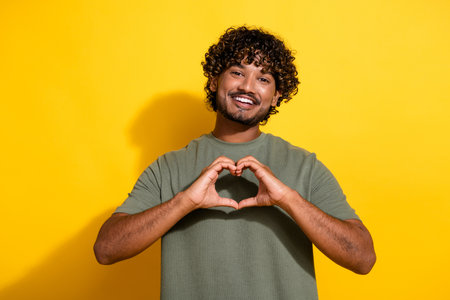 Photo of appreciative positive guy with wavy hair dressed khaki t-shirt showing heart symbol on chest isolated on yellow color backgroundの写真素材
