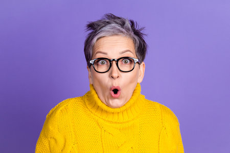 Surprised senior woman in yellow sweater with glasses against purple background