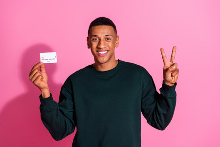 Photo of funky cool guy dressed pullover holding bank card showing v-sign emtpy space isolated pink...