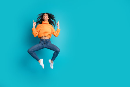 Full length photo of adorable lovely girl orange knitwear jumping high pointing up empty space isolated blue color backgroundの写真素材