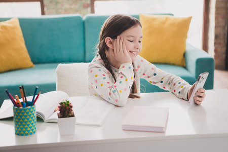 Photo of cute small schoolgirl study desk use smart phone wear pullover modern interior apartment indoorsの写真素材