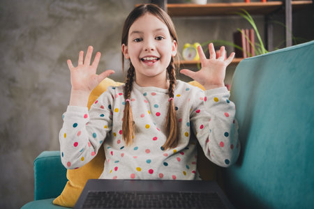 Photo of cute small schoolgirl web camera video call arms wave wear pullover modern interior apartment indoorsの写真素材