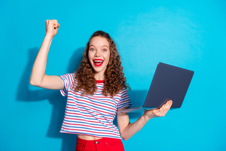 Excited young woman celebrates success with laptop against a vibrant blue background, expressing joy...